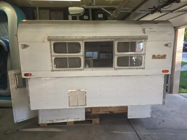 Alaskan camper exterior before restoration, with aged white siding and a broad bank of windows