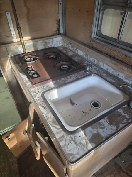Original camper galley with a worn sink counter, three-burner cooktop and unfinished plywood walls