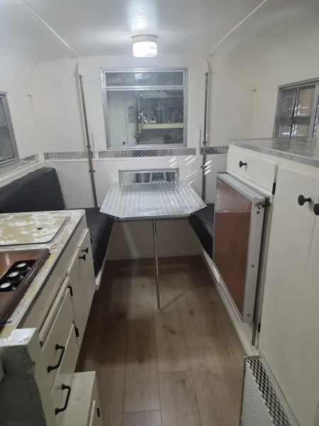 Refinished Alaskan camper interior with white cabinetry, a dinette table, black cushions and wood-look flooring