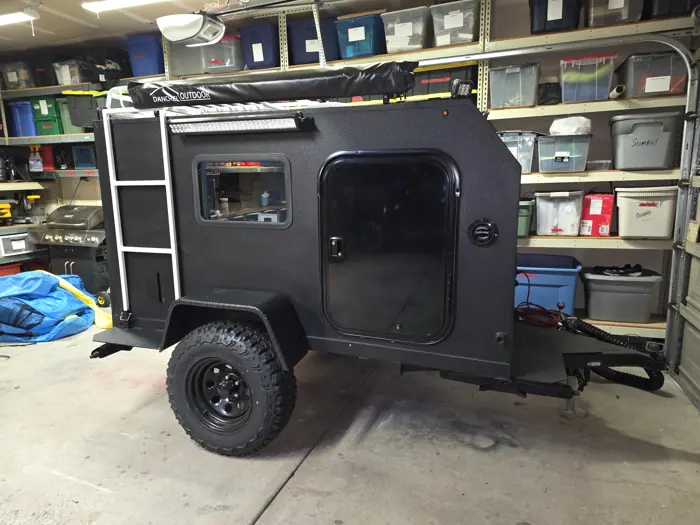 Black compact trailer with a side door, ladder, roof rack and off-road tires.