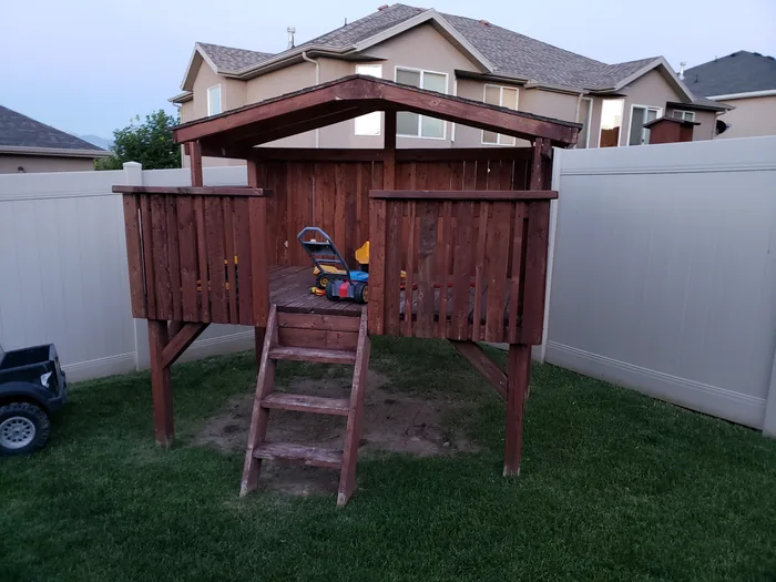 Completed elevated wood fort with a ladder, railings and shingled roof.